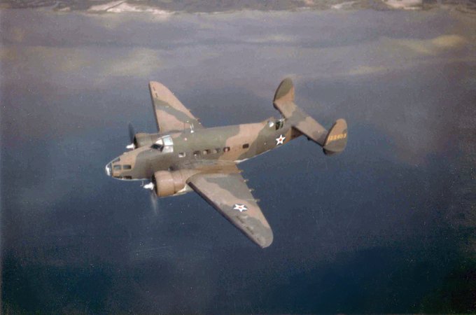 A vintage military twin-engine bomber plane with twin tail design flies over water, painted in camouflage with white star insignias.