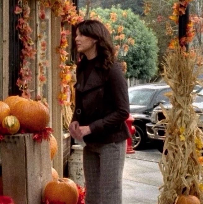 A woman in a dark coat stands outdoors near a display of pumpkins and autumn garlands, looking sideways in a scene from what appears to be a TV show.