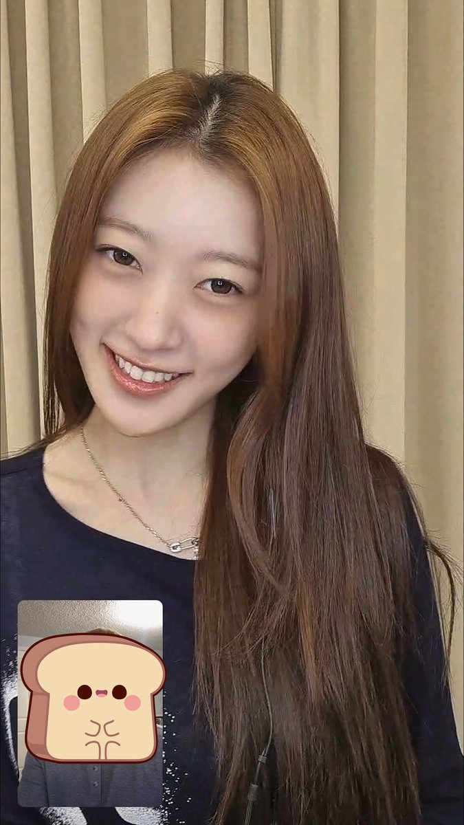 A smiling young woman with long brown hair wearing a dark blue top poses for a selfie video call, with a cartoon toast sticker partially covering her face.
