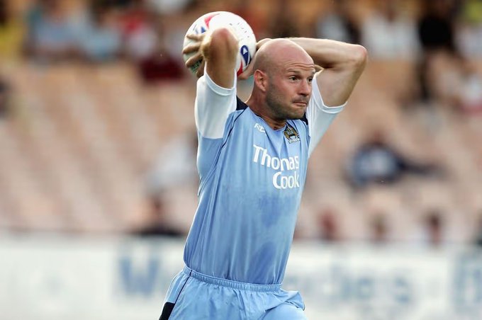A bald soccer player in a light blue 'Thomas Cook' jersey prepares to throw the ball into play with a stadium crowd blurred in the background.
