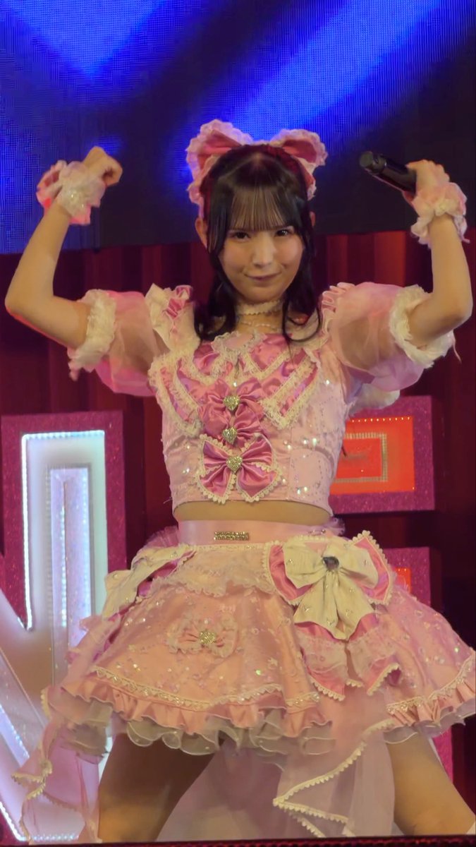 A young woman with dark hair wearing an elaborate pink frilly idol performance outfit with bows, posing on stage with a microphone and a matching cat-ear headband.