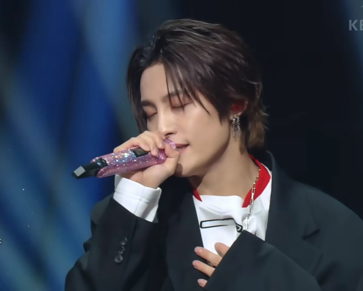 A performance shot of a K-pop idol singing with eyes closed while holding a sparkly pink microphone, wearing a black suit jacket over a white shirt with red accents and dangling earrings.