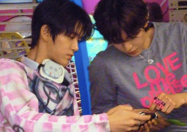 Two young men with dark hair look down at something together, one wearing a pink and white plaid shirt with headphones around his neck, the other in a gray 'LOVE' t-shirt with a necklace.