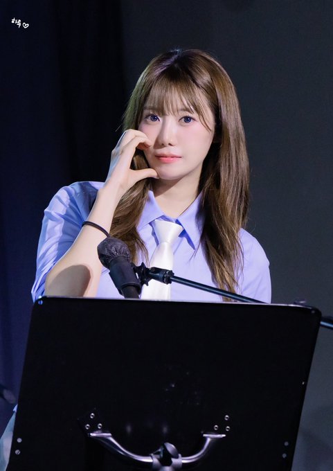 A young woman with long brown hair and bangs sits behind a music stand and microphone, resting her hand near her face, wearing a light blue collared shirt and white tie.