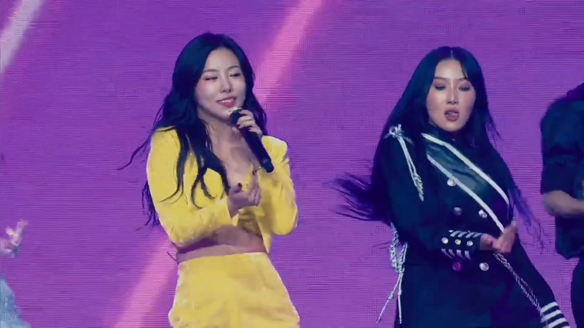 Two female performers dance on stage against a vibrant pink backdrop; the singer on the left wears a yellow outfit while the dancer on the right wears a black military-style jacket with chains.