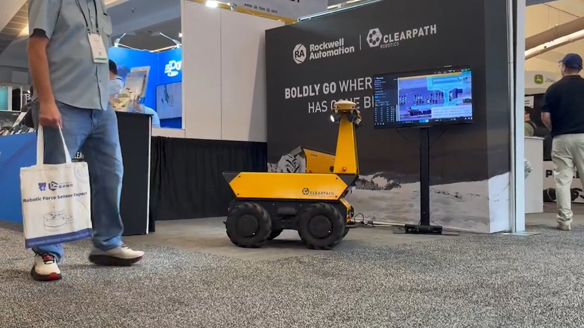 Trade show booth for Rockwell Automation and Clearpath Robotics with the slogan 'BOLDLY GO WHERE [NO ONE] HAS GONE B[EFORE]', displaying a yellow Clearpath mobile robot next to a person holding a 'Robotic Force Sensor Expert' tote bag.