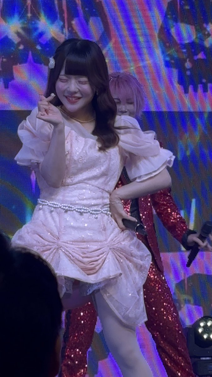 A young woman in a sparkly pink dress with a pearl belt poses on stage with a finger heart, backed by another performer in a red sequin outfit.