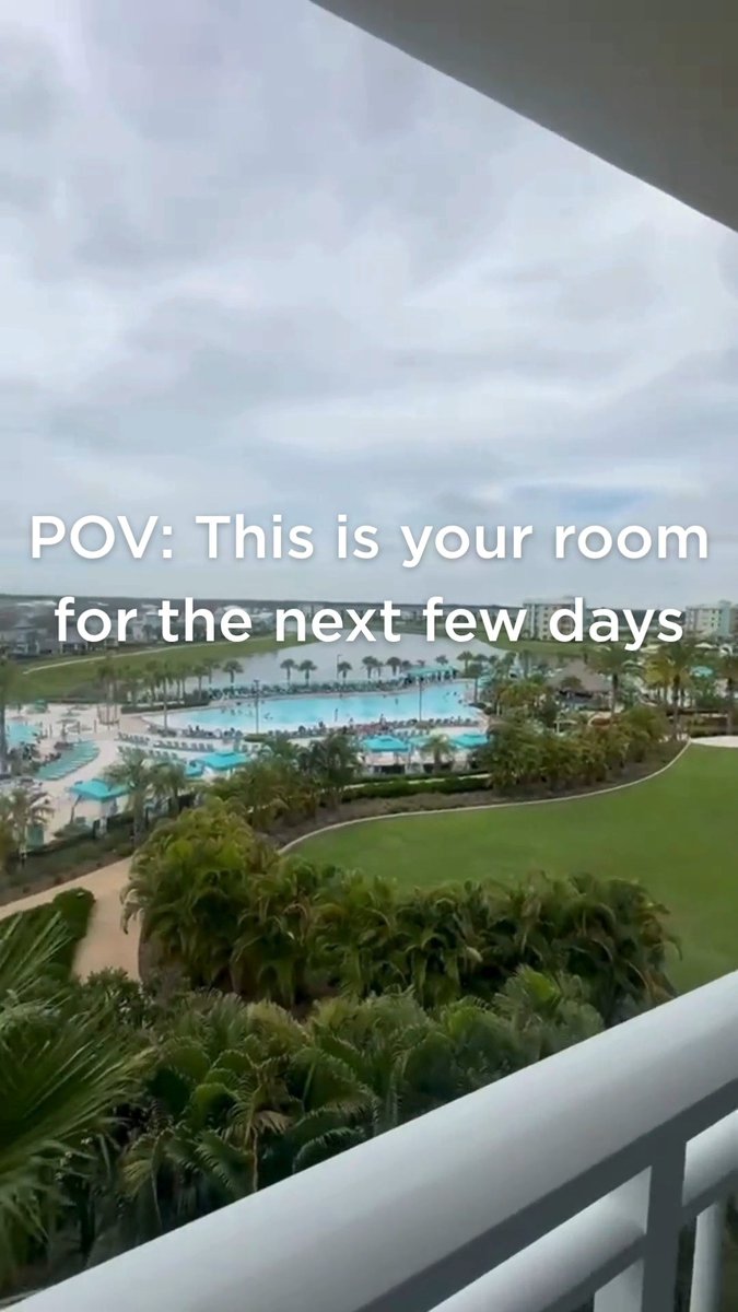 A view from a hotel balcony overlooks a resort pool, palm trees, and a lake, with text overlay reading 'POV: This is your room for the next few days.'