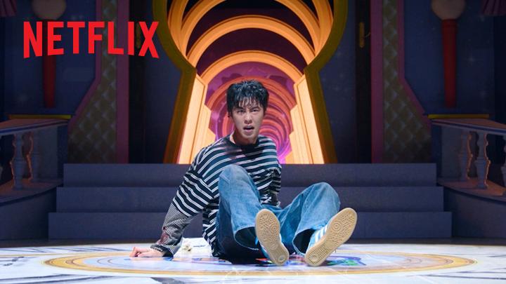 A young man sits on the floor of a brightly lit, ornate set with the "NETFLIX" logo visible in the top left corner, looking startled as colorful items spill out around him. The image aligns with the post's theme, depicting Lee Jae-wook in a moment of shock as he contemplates the ominous job offer described in the caption.