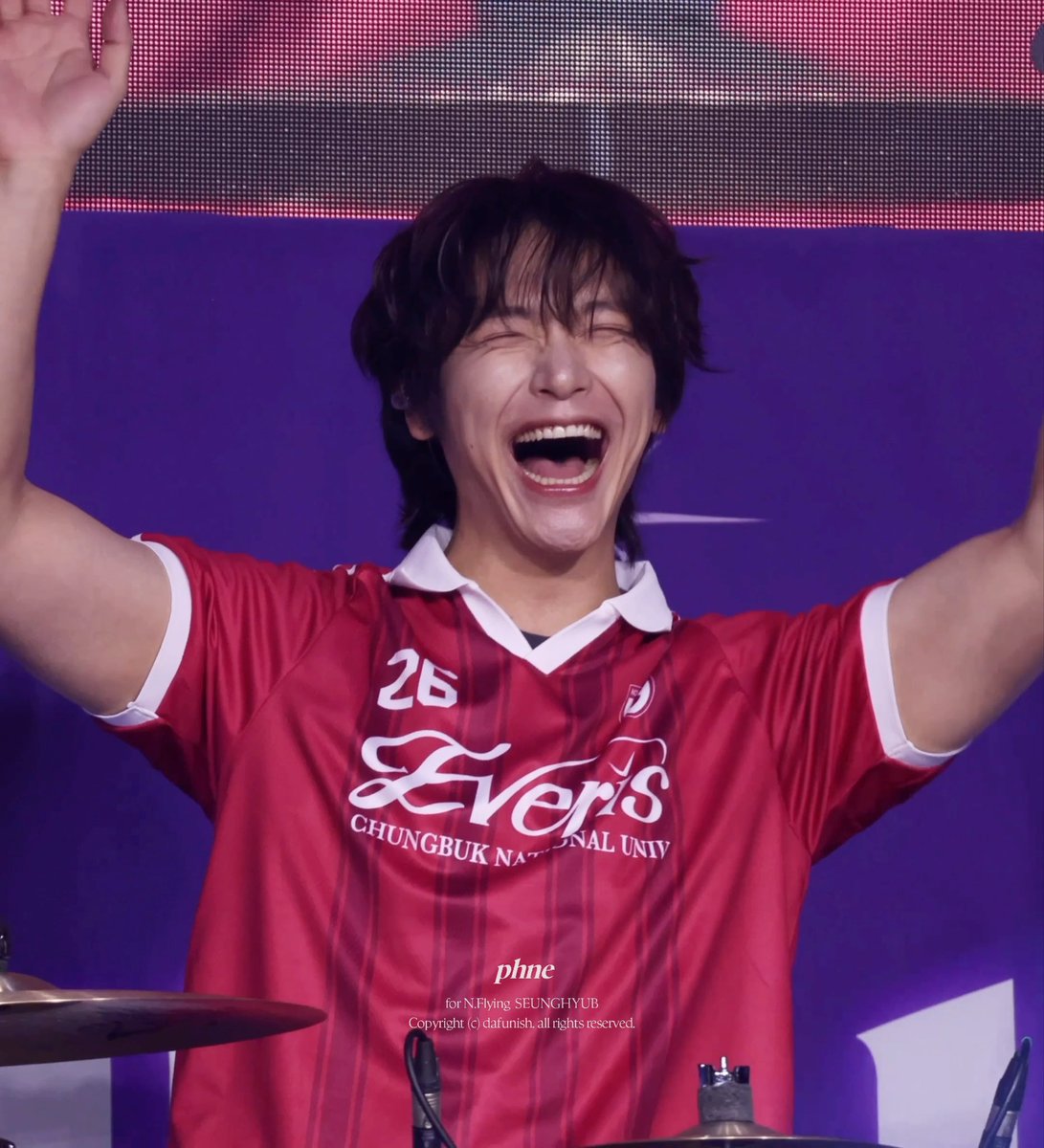 A young man with dark hair enthusiastically cheering with arms raised while wearing a red '26 Evers Chungbuk National University' jersey on stage.