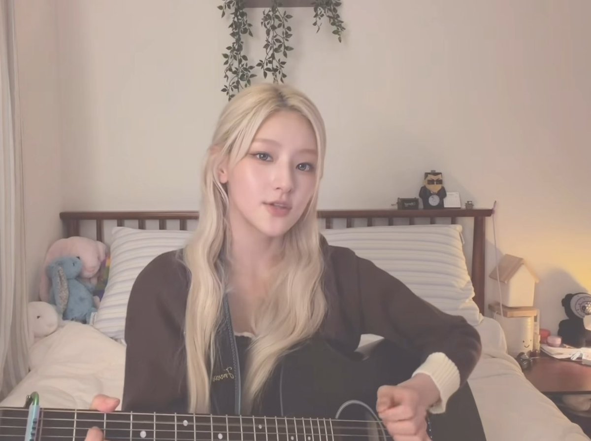A young woman with long blonde hair sitting on a bed and playing an acoustic guitar in a softly lit room.