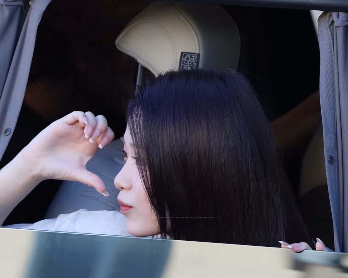 A young woman with long dark hair sits inside a luxury car with a Rolls-Royce headrest emblem, making a heart gesture with her hand; faint text reads 'BTSEGEN MAGLIE K-CULTURE FESTIVAL ON THE WAY HOME'.