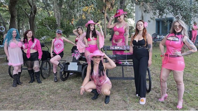 A group of women pose together outdoors in pink-themed costumes and cowboy hats, surrounded by trees and a picnic table.