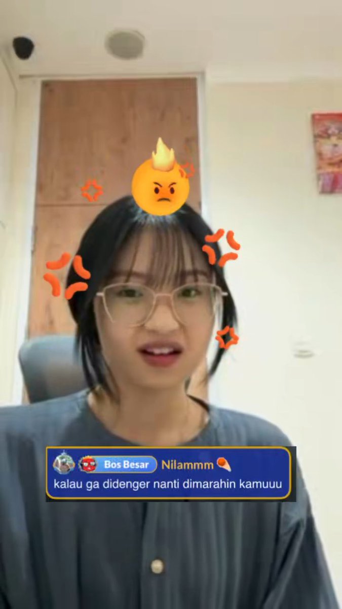 A screenshot of a video shows a young woman with glasses and a digital angry emoji filter, with a comment overlay reading 'Bos Besar Nilammm kalau ga didenger nanti dimarahin kamuuu'.
