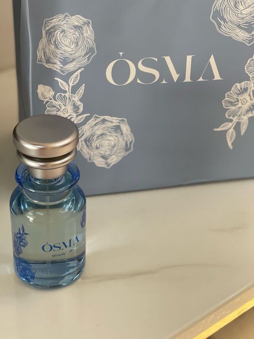 A small blue perfume bottle labeled 'OSMA Woody W' sits in front of its matching blue packaging box, which features floral illustrations and the brand name 'OSMA'.