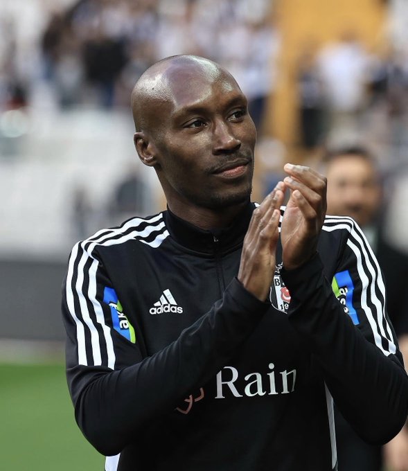 Atiba Hutchinson, wearing a black Beşiktaş training jacket with the Adidas logo and "Rain" sponsor text, is shown clapping his hands in what appears to be a stadium setting, likely shown in context as he comments on fellow players Dusan Vlahovic and Trossard.