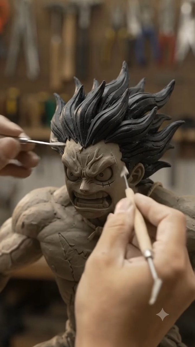 An artist's hands using sculpting tools to detail the face of a fierce clay sculpture of a spiky-haired, muscular character.
