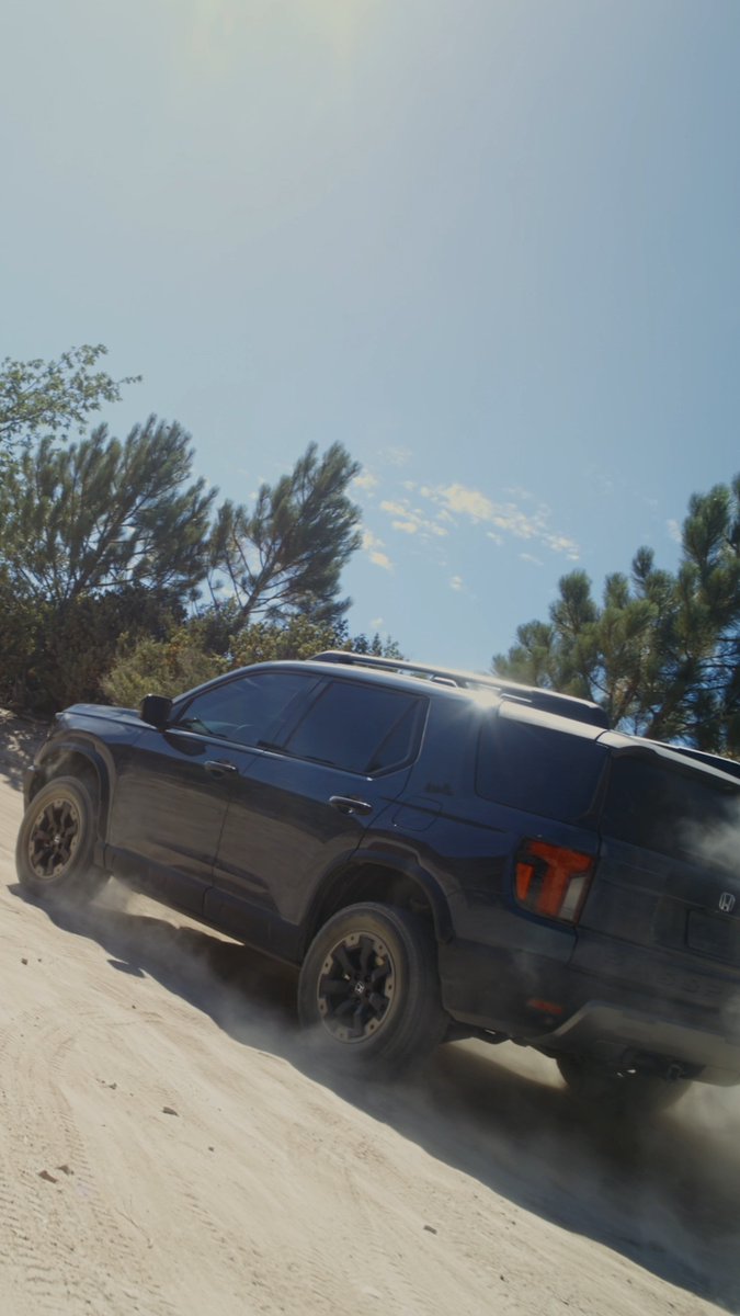 A dark blue Honda Passport TrailSport SUV kicks up dust as it powers through sandy off-road terrain under a bright blue sky, showcasing its rugged capability. The image visually reinforces the post's message of enhanced ground clearance, capability, and adventure-ready performance for the 2027 model.