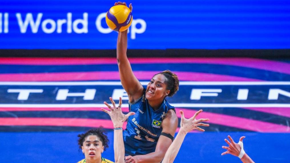 Ana Cristina, wearing the iconic blue Fenerbahçe jersey with the Brazilian flag, spikes the ball over two Brazilian defenders during a Volleyball World Cup match, illustrating her starring role ahead of her sixth season with the Turkish club.