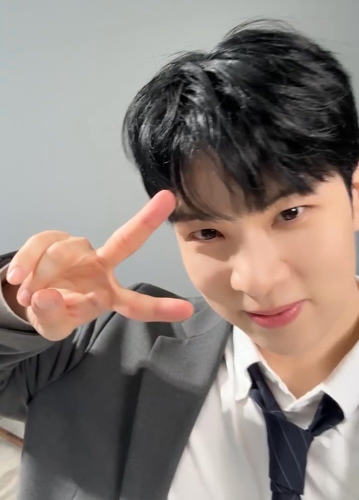 A close-up selfie of a young man with dark hair wearing a dark suit and white shirt, smiling and making a hand gesture near his face.