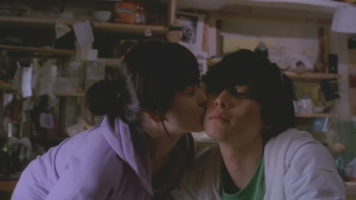 Film still showing a woman in a lavender robe kissing a man wearing sunglasses and a white jacket on the cheek, set in what appears to be a cluttered store or shop.