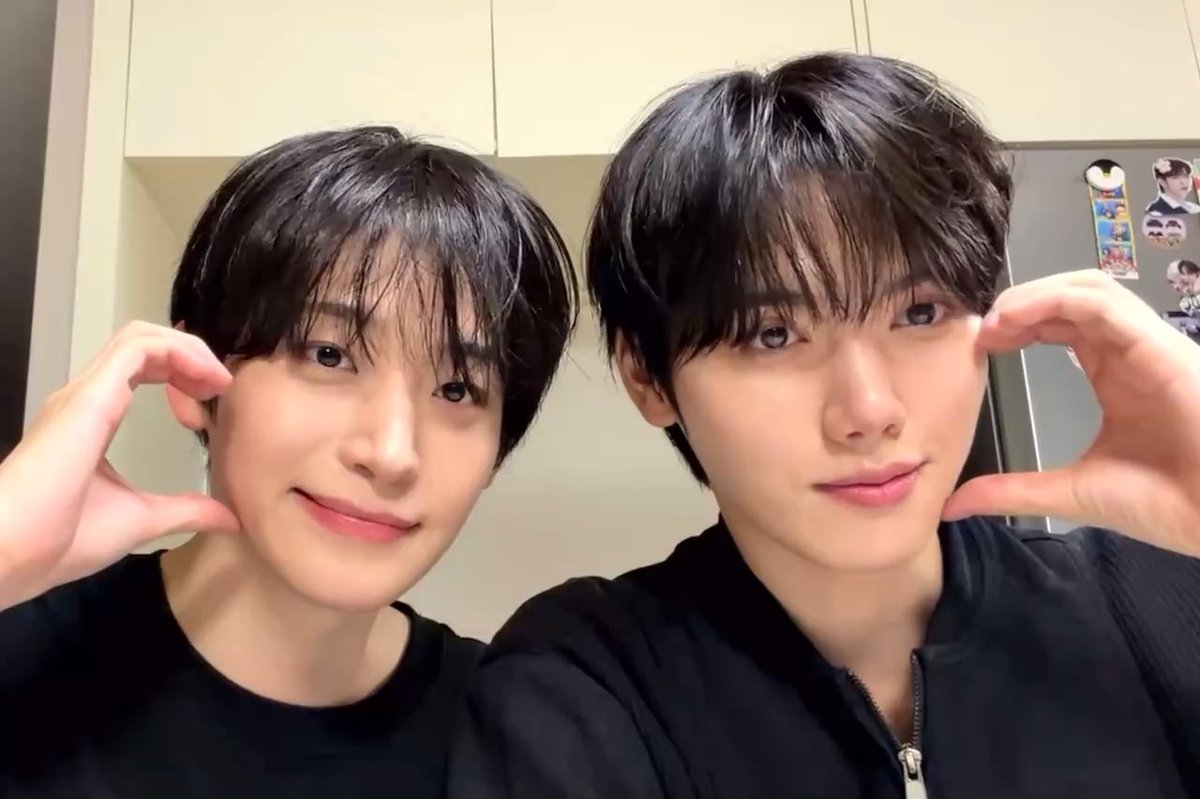 Two young men with dark hair taking a selfie, making a frame gesture with their hands near their faces in what appears to be a kitchen.
