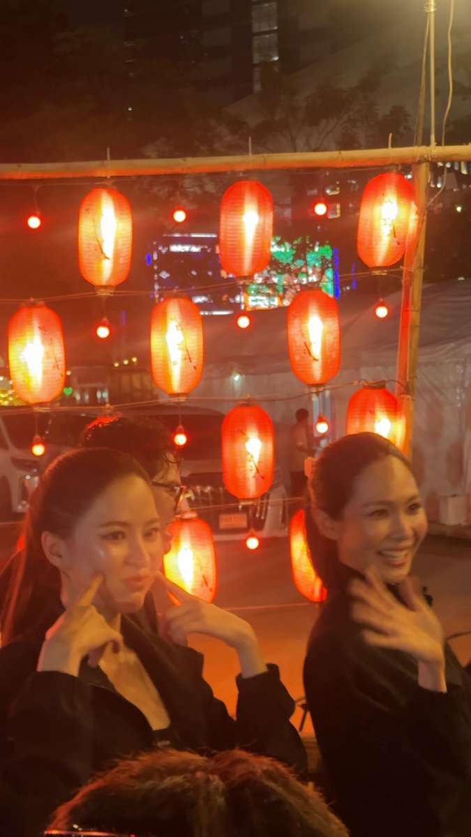 Two women pose in front of a row of glowing red Japanese-style paper lanterns at a nighttime outdoor event.