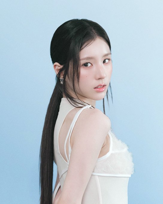 A portrait of a young woman with long dark hair tied back, wearing a white top and looking over her shoulder against a light blue background.