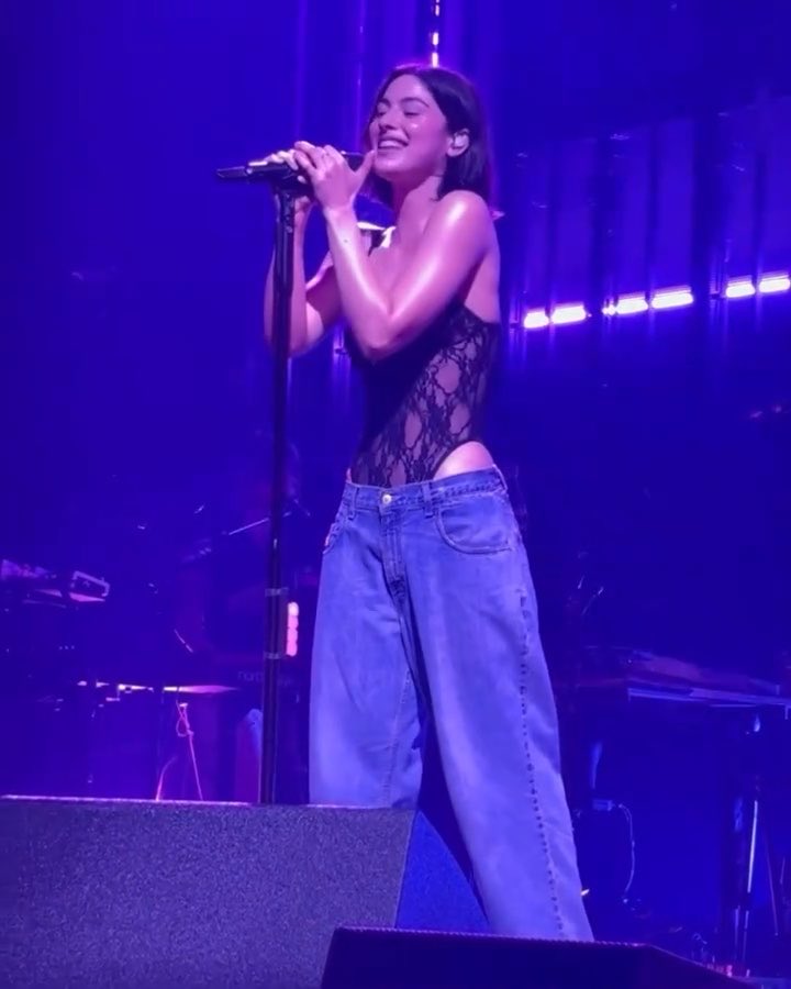 A young woman passionately sings into a microphone on a concert stage bathed in blue lighting, wearing a black snakeskin-patterned top and oversized jeans. The caption "take me back to this feeling" perfectly captures the euphoric, immersive moment of being lost in the music under the glowing stage lights.