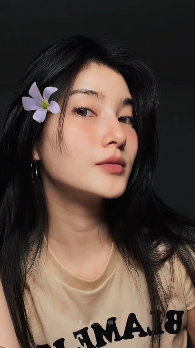 A portrait of a woman with long black hair and a light purple flower tucked behind her ear, wearing a beige sleeveless top with the letters 'PHUPE FAMLB' visible, gazing to the side.