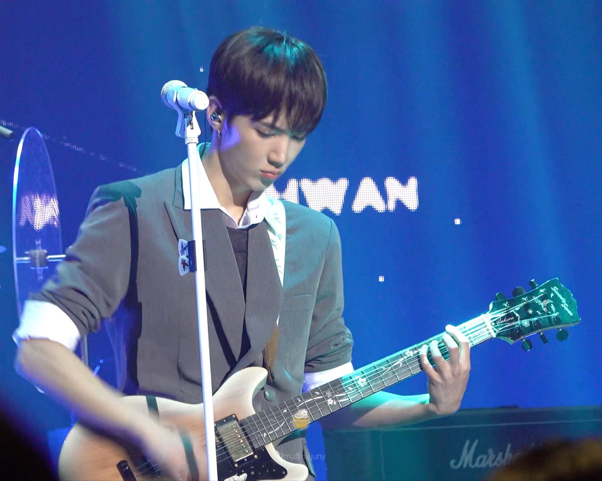 A young male musician performs on stage in a gray blazer over a white shirt, playing a white electric guitar in front of a microphone. The backdrop shows partial text reading '...HYAN' and a Marshall amplifier is visible.