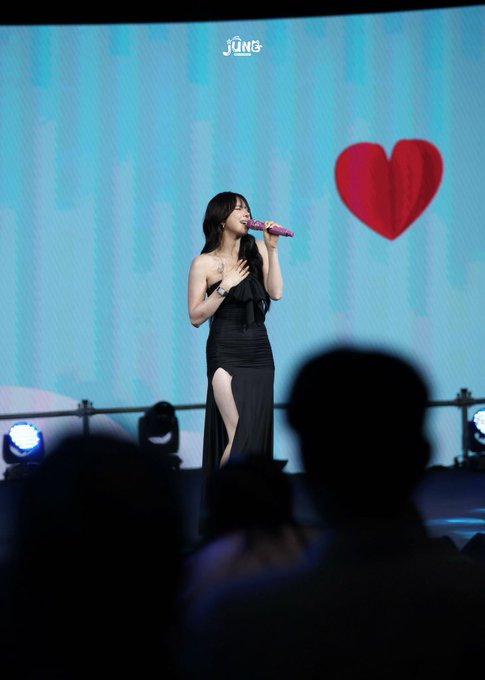 A female singer in a black dress performs on stage, holding a pink microphone, with a large red heart displayed on the screen behind her.