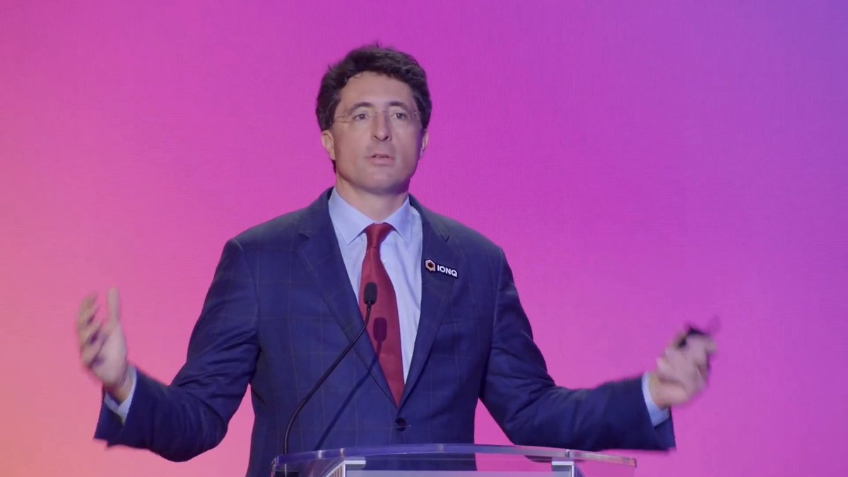 CEO Niccolo DeMasi, wearing a suit featuring an "IONQ" lapel pin, delivers a keynote address on a stage with a vibrant pink and purple gradient backdrop. His animated hand gestures convey the energetic presentation of IonQ's full-stack quantum strategy at the Quantum World Congress.