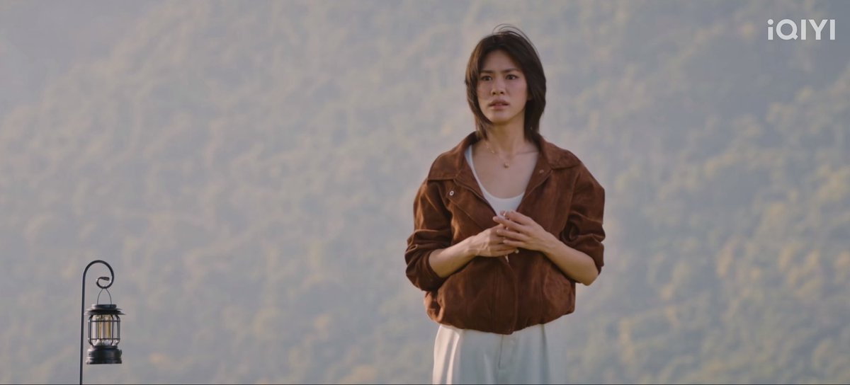 A still from an iQIYI video showing a woman with short hair wearing a brown suede jacket and white top, standing near a lantern with a blurry natural background.