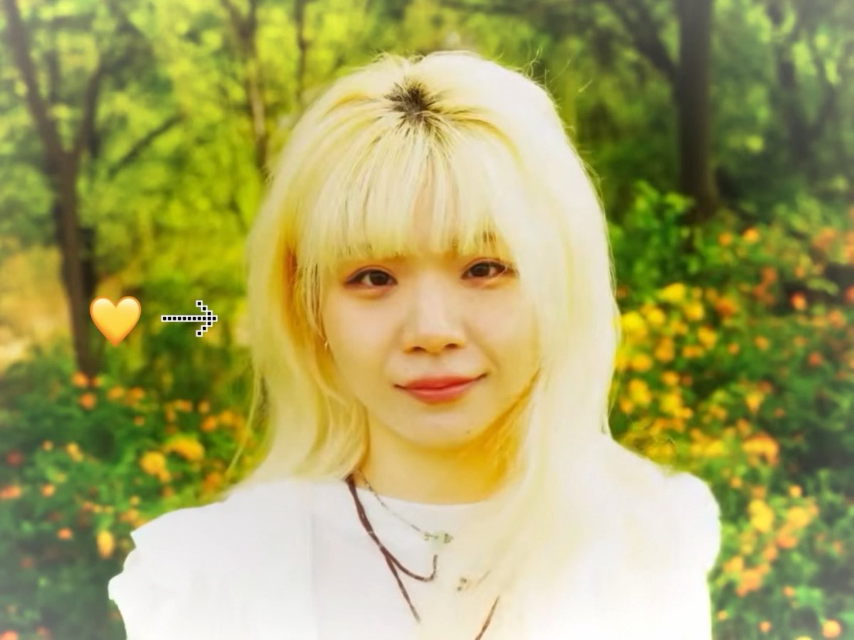 A young woman with blonde hair and bangs smiles softly, photographed in a sunlit garden with yellow flowers; an orange heart emoji and arrow graphic appear on the image.