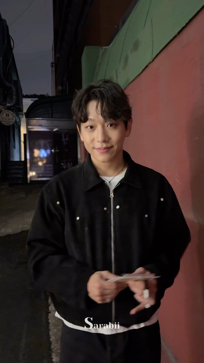 A young man with dark wavy hair stands in an alleyway at night in a black zip-up jacket, with the text "Sarabii" visible at the bottom of the image.