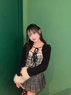 A young woman with long dark hair poses against a green wall, wearing a plaid dress with a black cardigan featuring fluffy white trim.