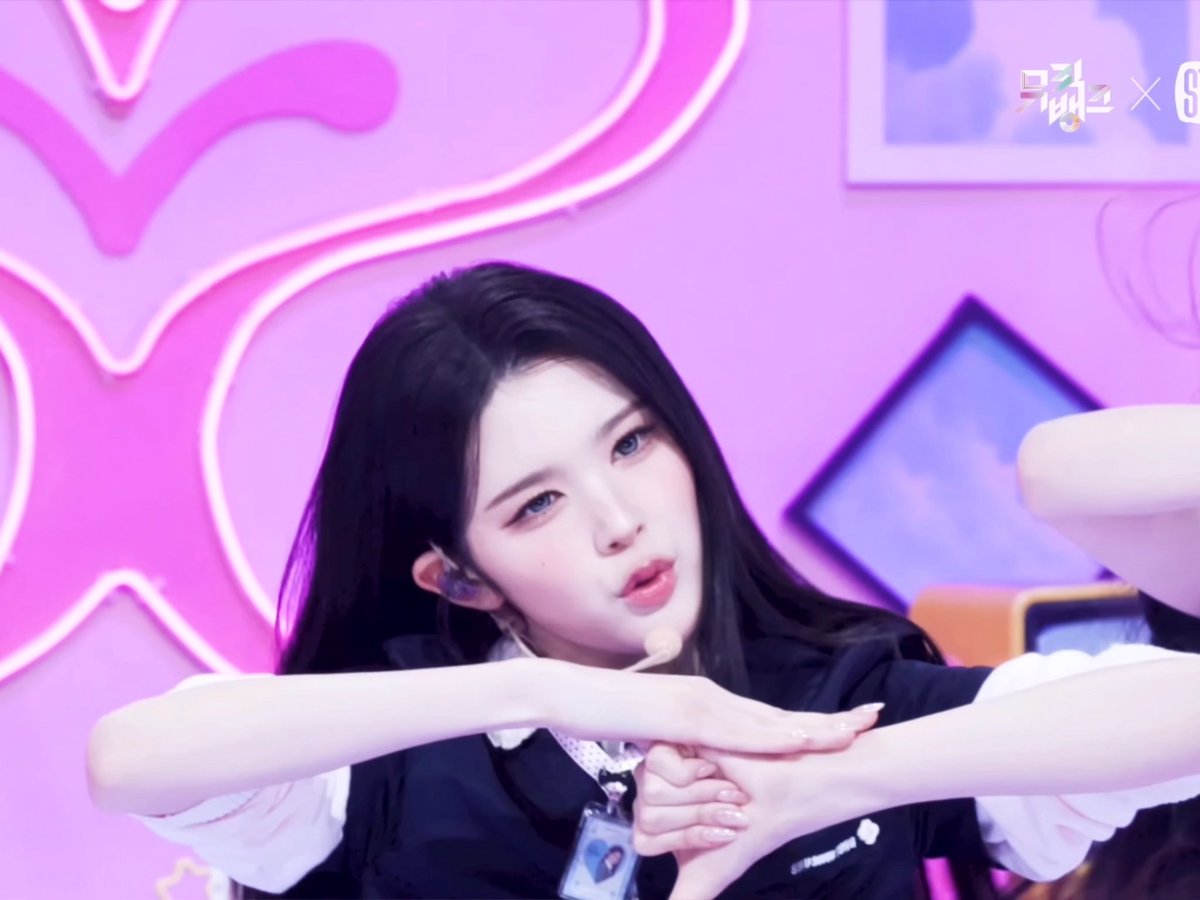 A young woman with long dark hair and striking blue eyes performs a dance move on a stage with a bright pink background featuring star and neon light designs.