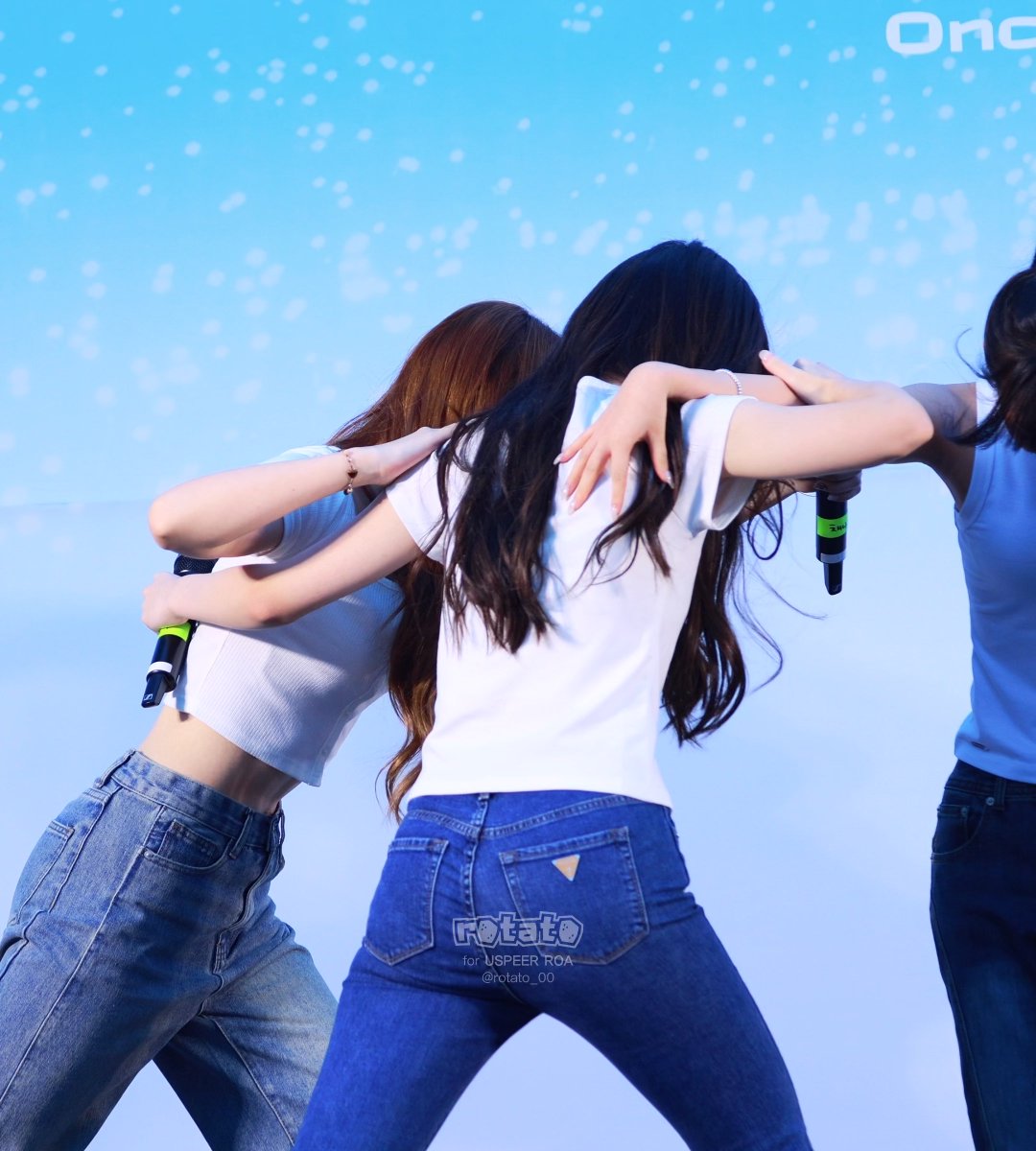 Three performers in white and blue tops with jeans pose on stage with arms around each other, holding microphones, with a blue starry background and partial text 'On' visible.