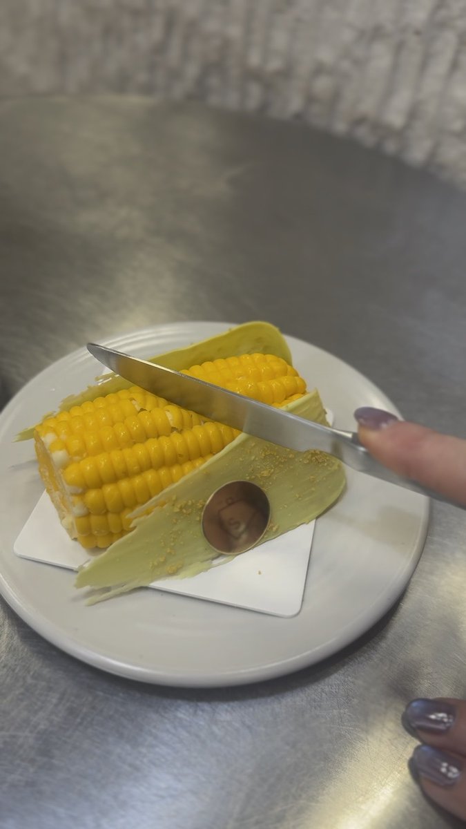 A first-person view of someone holding a knife to cut a cob of corn on the cob, served on a white plate with the husk pulled back.