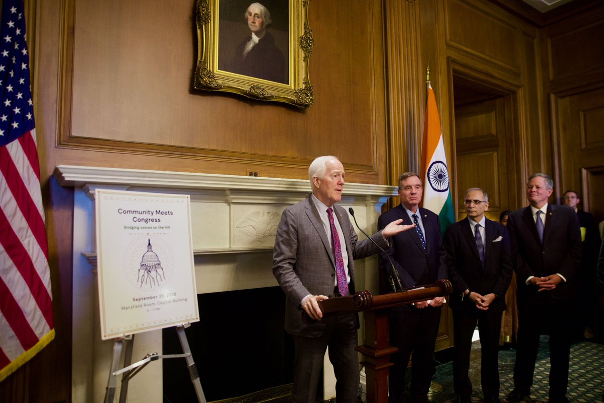 A man speaks at a wooden lectern in a formal, wood-paneled room while several men stand behind him. U.S. and Indian flags, a framed portrait, and a presentation board are visible. The board contains text.
