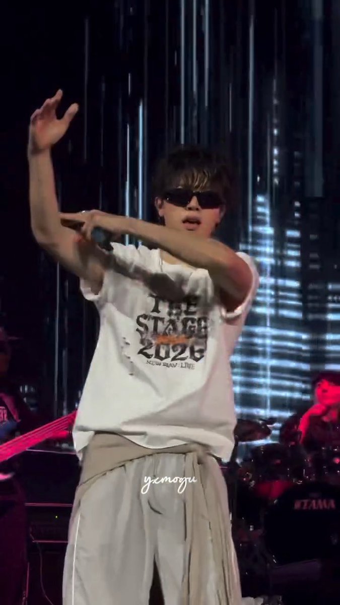 A male performer on stage wearing sunglasses and a distressed white t-shirt that reads "THE STAGE 2026," with a drum kit and a "TAMA" logo visible in the background.