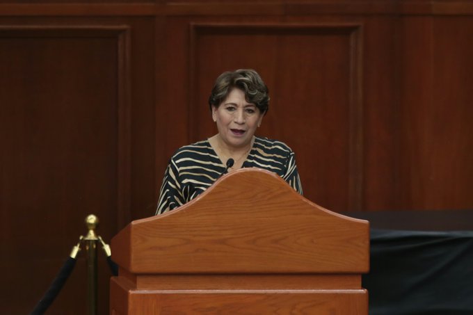 A woman with short brown hair stands at a wooden podium speaking into a microphone, set against a dark wood-paneled wall.