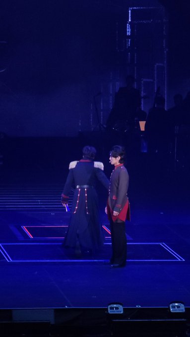Two performers in dark military-style costumes with red accents stand facing each other on a stage lit in deep blue.