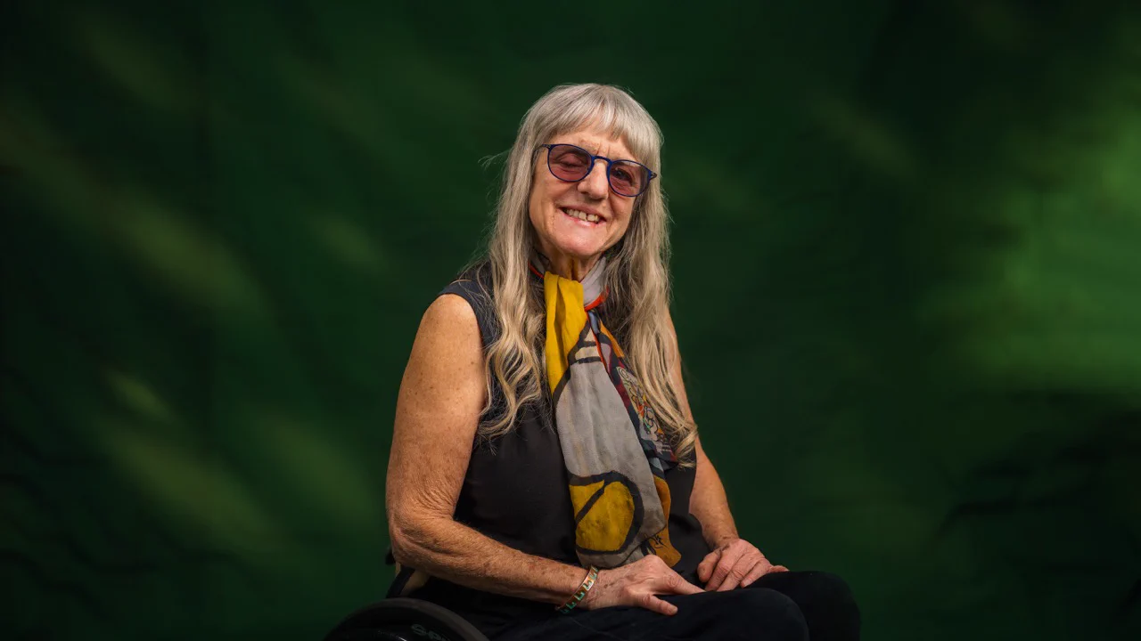 Individual sits in a wheelchair against a green and yellow background. The person is smiling, wearing a bright scarf and glasses.