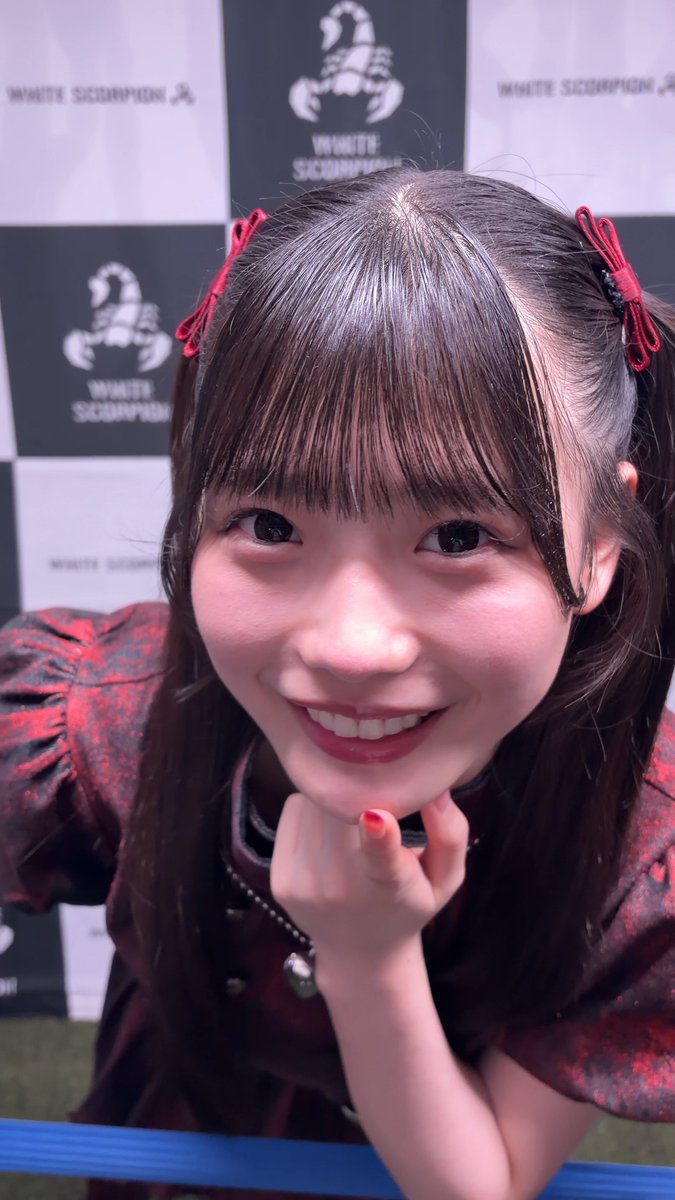 A smiling person with dark hair styled in pigtails with red ribbons, resting their chin on their hand in front of a backdrop with 'WHITE SCORPION' logos.