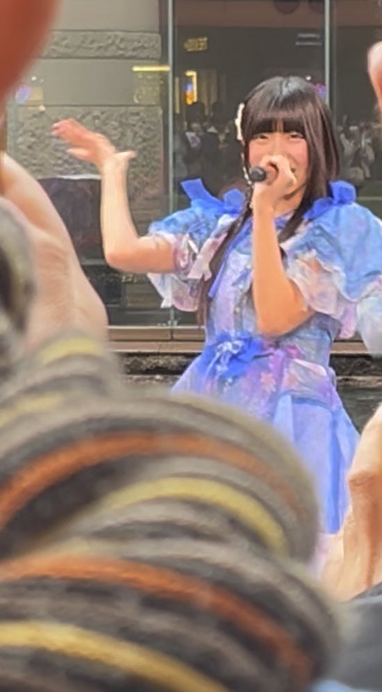 A performer in a blue floral dress sings into a microphone on stage at what appears to be an outdoor event.