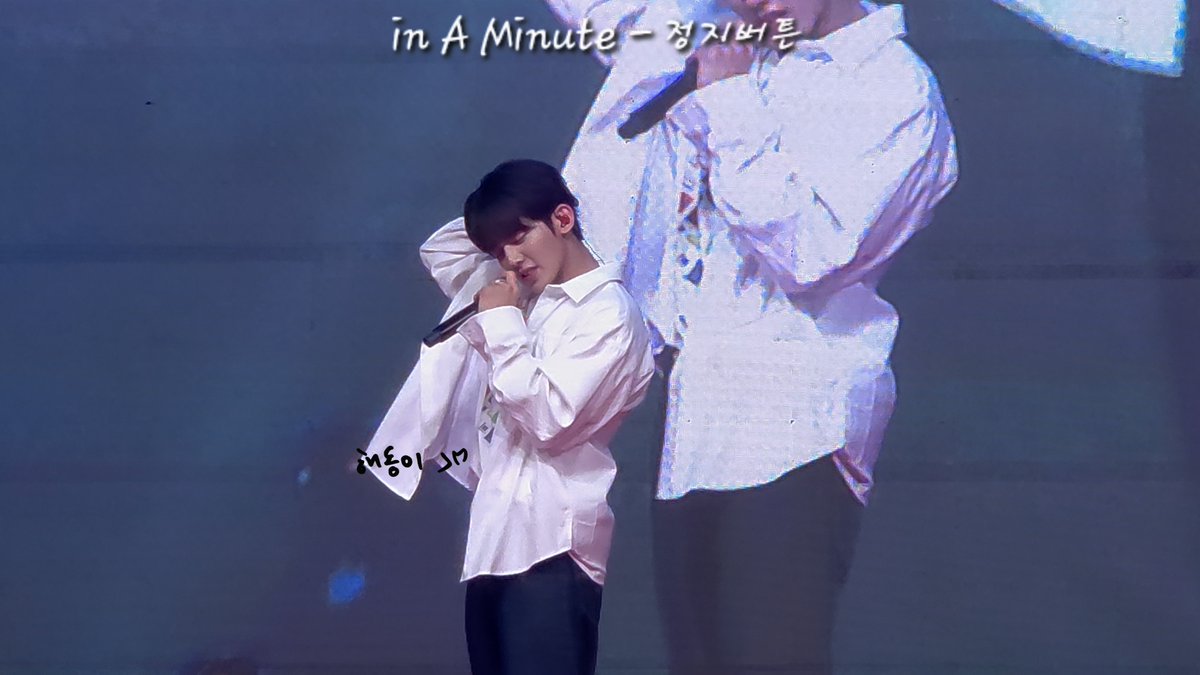 A man in a white shirt performs on stage holding a microphone, with the text 'in A Minute - 정치범들' visible at the top and a Korean annotation near his hands.