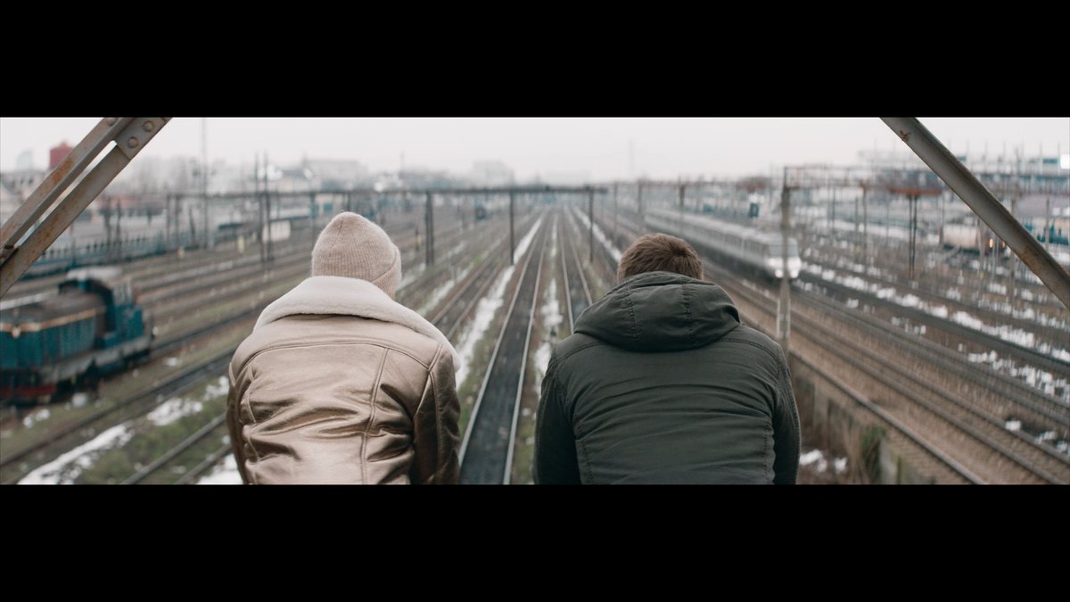 Two figures sit side by side on an overpass overlooking a sprawling rail yard, their backs to the camera as they gaze down at the converging tracks below. The bleak, wintry industrial setting evokes the gritty tension of the film's premise—a descent into a criminal underworld in search of his sister.