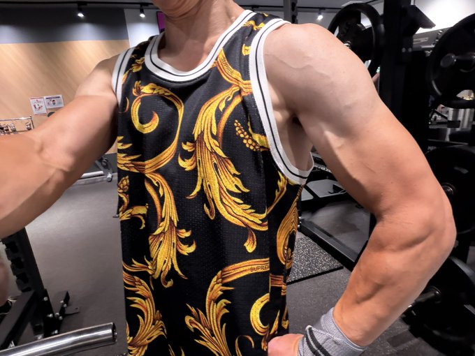 A muscular man in a gym wearing a black and gold baroque-patterned tank top and wrist wraps.